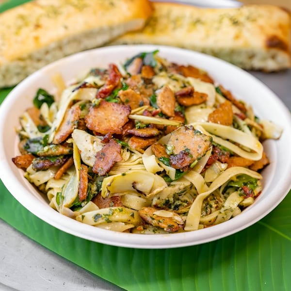 A white bowl of fettuccine pasta with sausage, spinach, and sun-dried tomatoes, with garlic bread in the background.