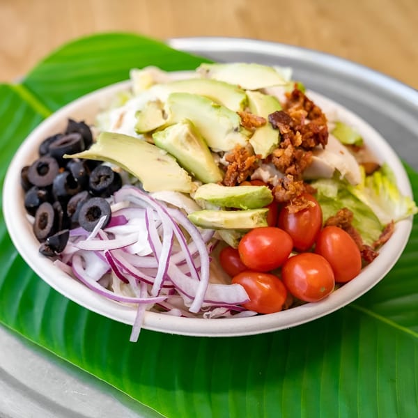 A vibrant salad with avocado, olives, red onions, cherry tomatoes, and bacon bits in a white bowl.