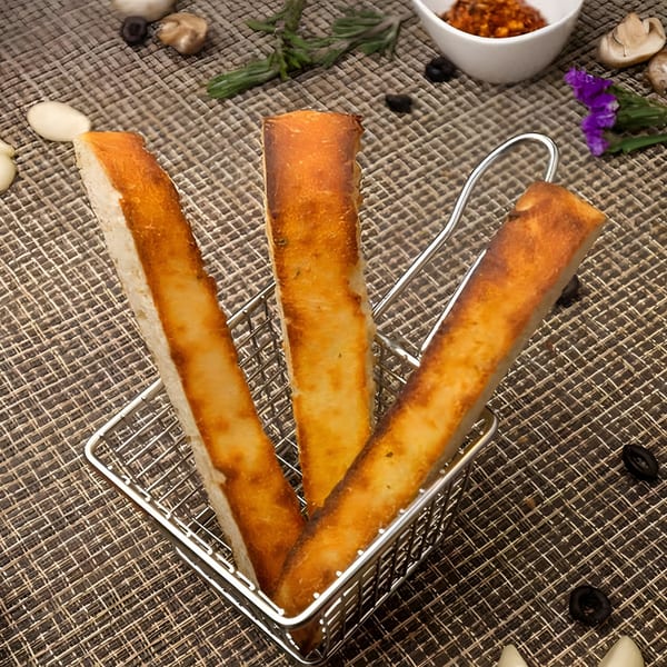 Three golden-brown breadsticks in a metal basket, with garlic, olives, and chili flakes scattered around.
